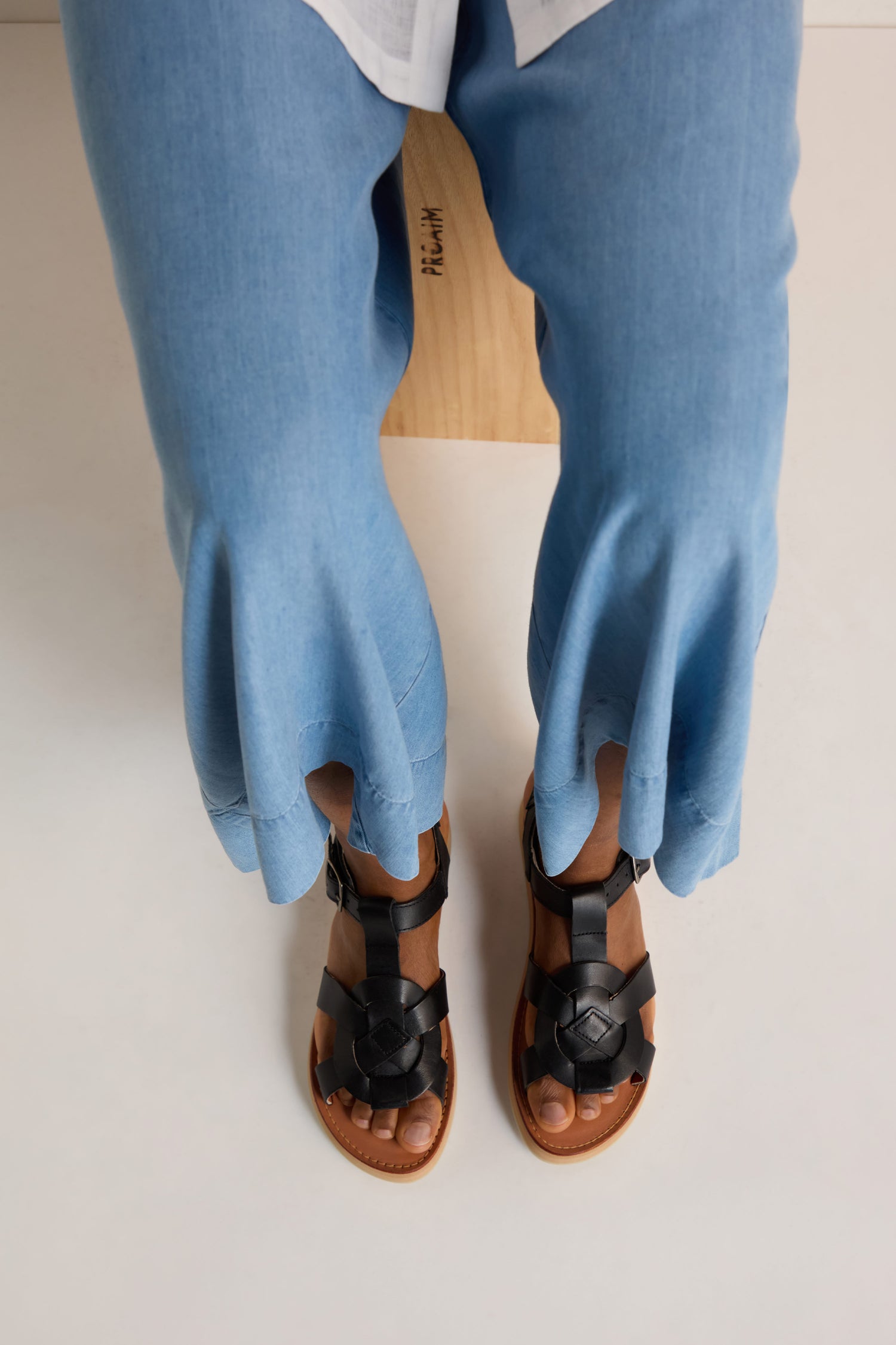 Person wearing light blue flared pants and Tulum Leather Sandals, seated on a wooden stool and photographed from above.