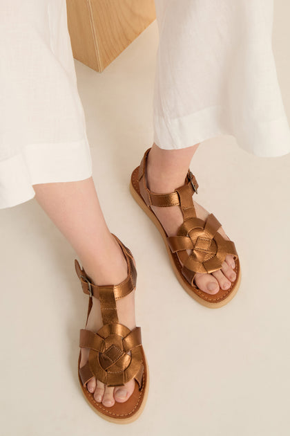 Person wearing white pants and stylish Tulum Leather Sandals featuring a woven leather design, standing on a light surface.