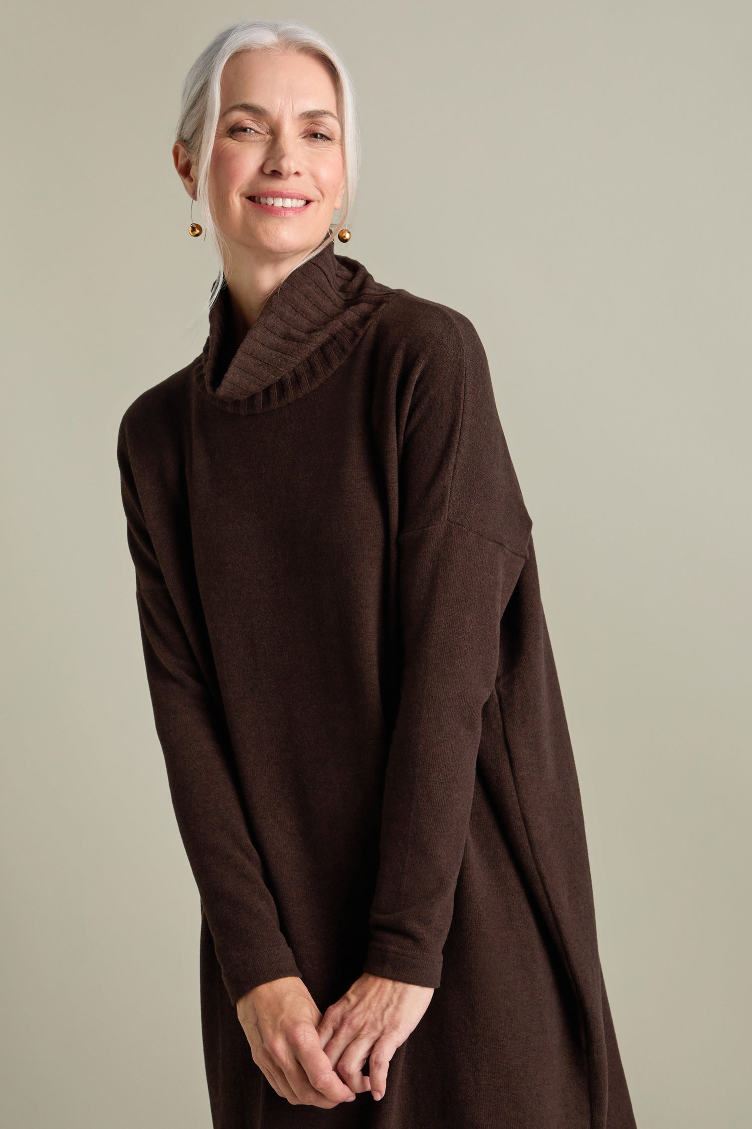 A woman with gray hair is wearing a long-sleeved, dark brown Allai Rib Neck Soft Jersey Dress, smiling softly against a plain background. This versatile addition to your wardrobe features a textured collar that adds an elegant touch.