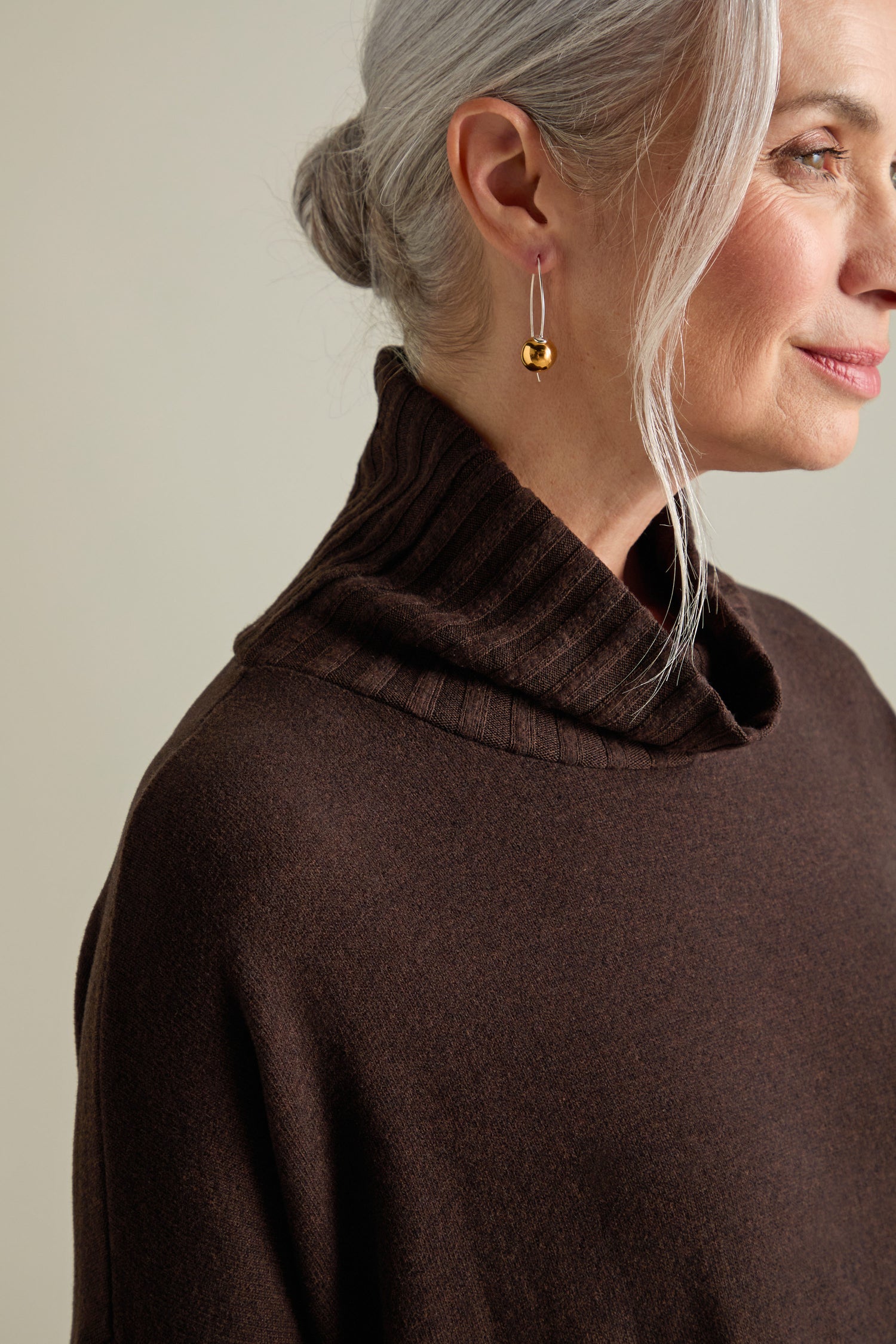 A woman with gray hair tied back, wearing gold earrings and a luxurious Allai Rib Neck Soft Jersey Dress, is shown in profile.