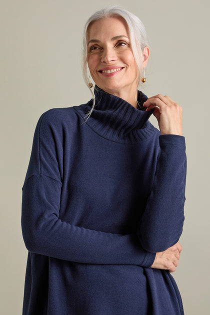 A woman with grey hair in a loose ponytail is smiling and wearing the Allai Rib Neck Soft Jersey Dress, a versatile addition to your wardrobe. She is holding the collar with one hand, against a plain beige background.