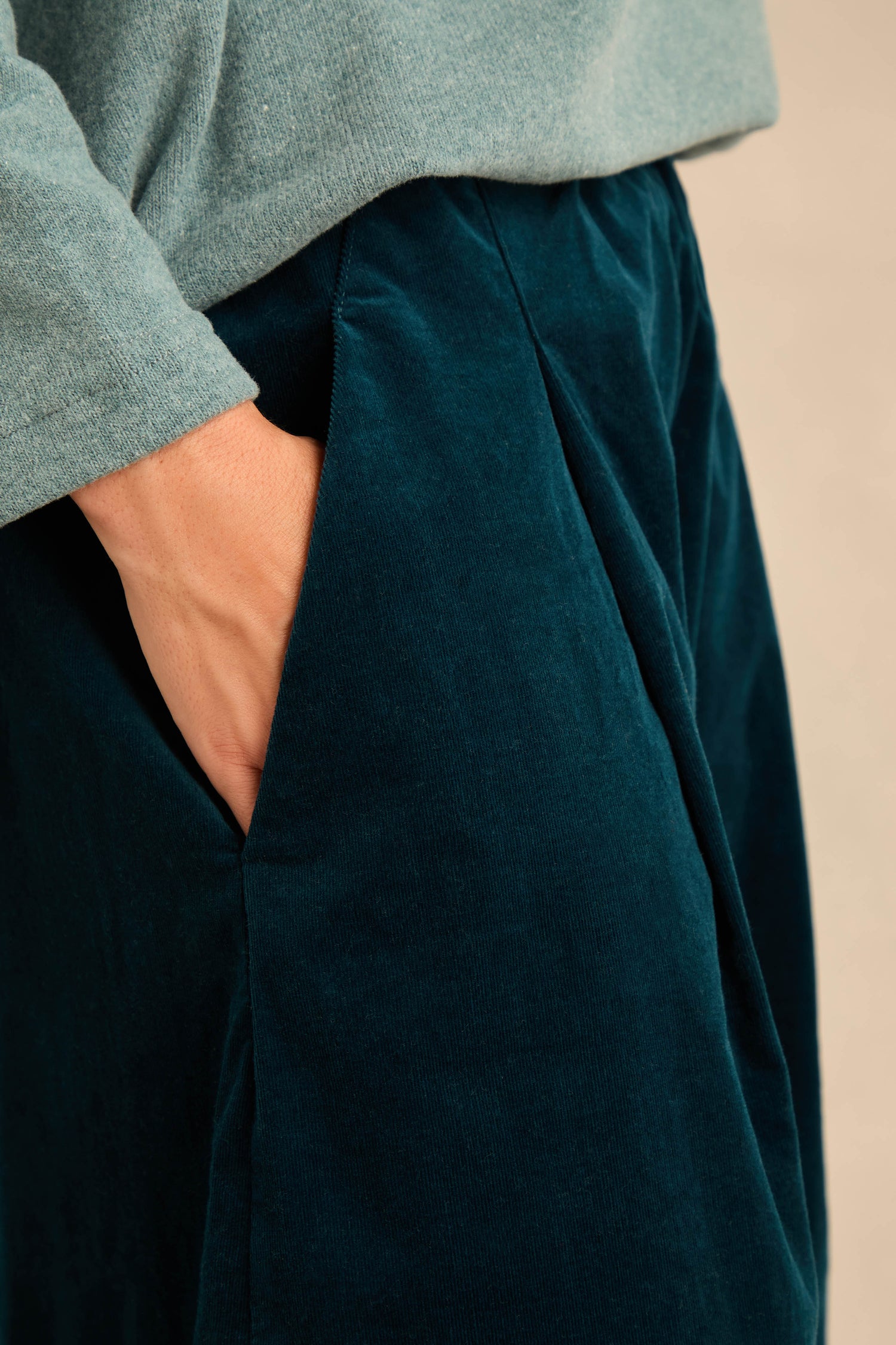 A person wearing a light green long-sleeve shirt and Stretch Velvet Pleat Front Trousers in dark teal stands with one hand casually placed in their pants pocket.