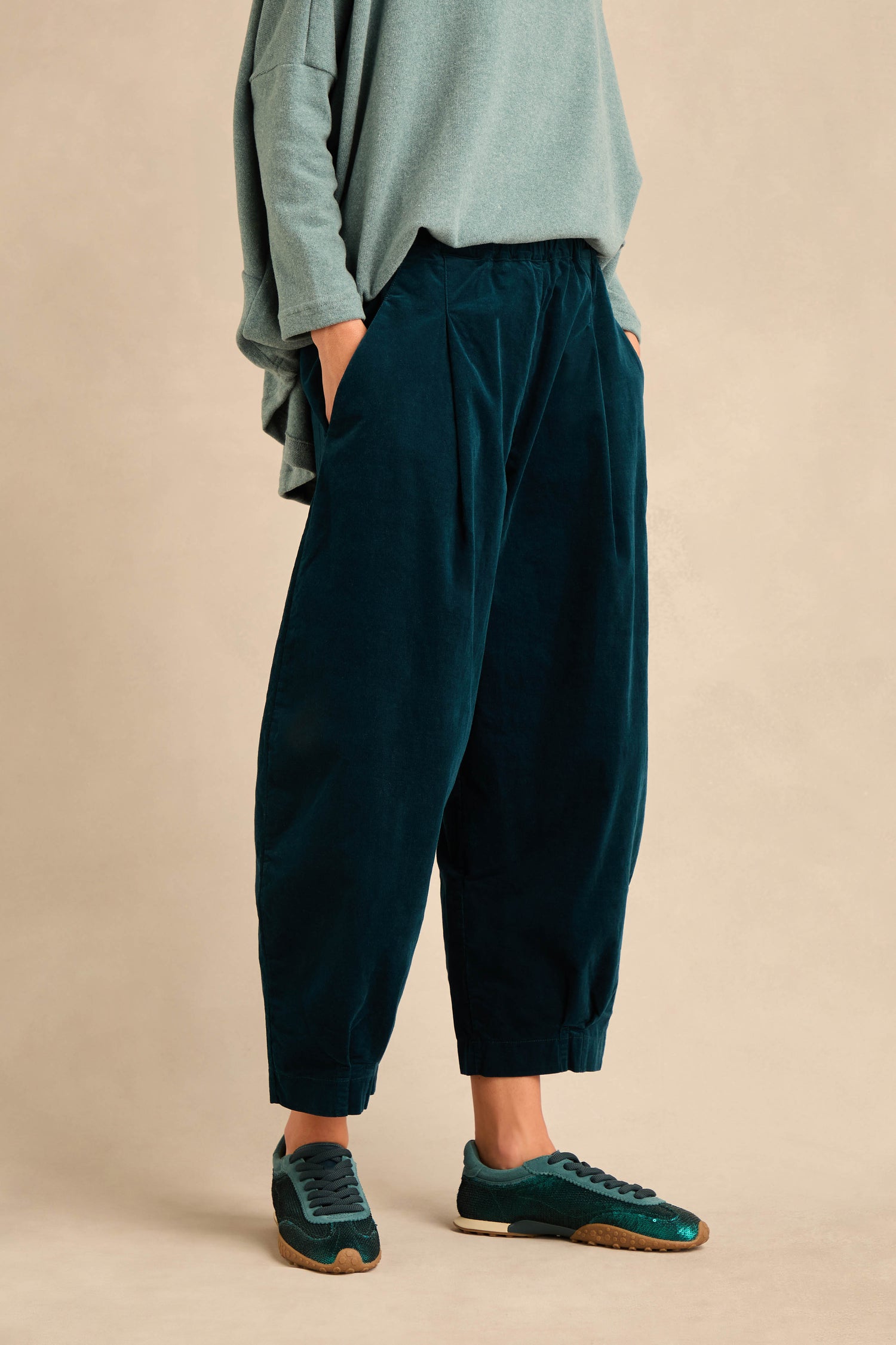 Wearing Stretch Velvet Pleat Front Trousers in teal, a light green oversized top, and matching teal sneakers, this person stands with hands in pockets against a plain beige background.