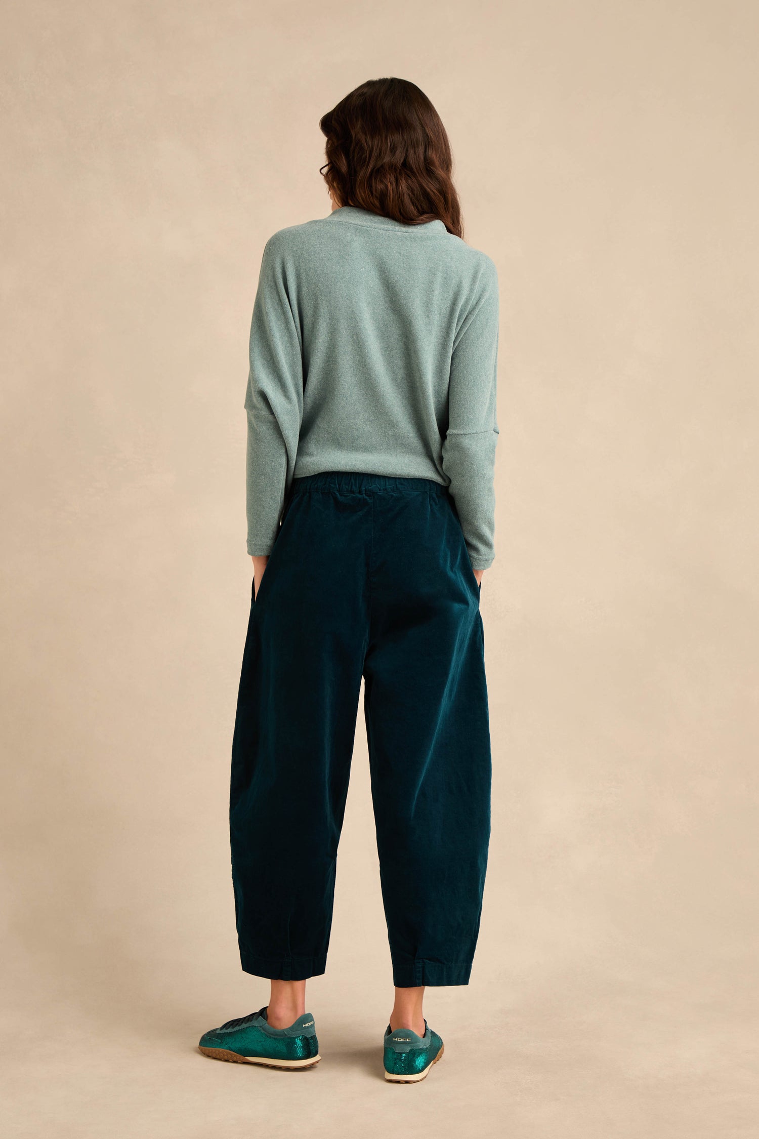 A person with long brown hair stands facing away, wearing a light blue sweater paired with Stretch Velvet Pleat Front Trousers in dark teal and matching teal shoes against a plain beige background.