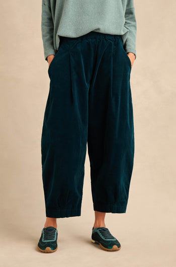 A person wearing Stretch Velvet Pleat Front Trousers in dark teal cotton velvet, paired with a light teal long-sleeve top and green sneakers, stands with hands in pockets against a beige background.