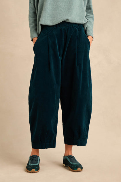 A person wearing Stretch Velvet Pleat Front Trousers in dark teal cotton velvet, paired with a light teal long-sleeve top and green sneakers, stands with hands in pockets against a beige background.