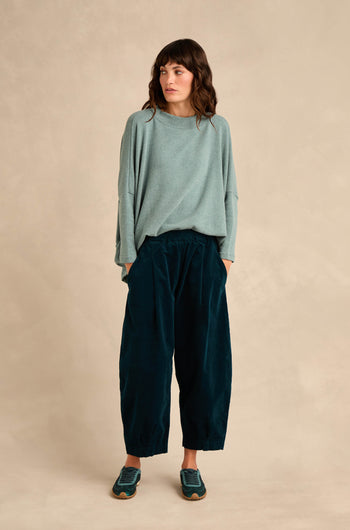 A person stands against a beige background wearing a loose light blue sweater, Stretch Velvet Pleat Front Trousers in dark blue, and blue sneakers, with hands in pockets and looking slightly to the side.