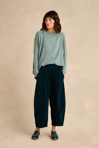 A person stands against a beige background wearing a loose light blue sweater, Stretch Velvet Pleat Front Trousers in dark blue, and blue sneakers, with hands in pockets and looking slightly to the side.