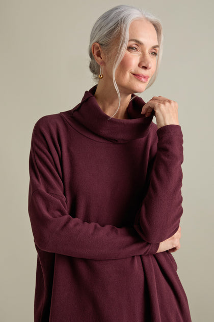 An older woman with gray hair tied back is wearing a long-sleeve, maroon Bill Roll Neck Soft Jersey Top and small gold hoop earrings. She stands against a neutral background, looking off to the side.