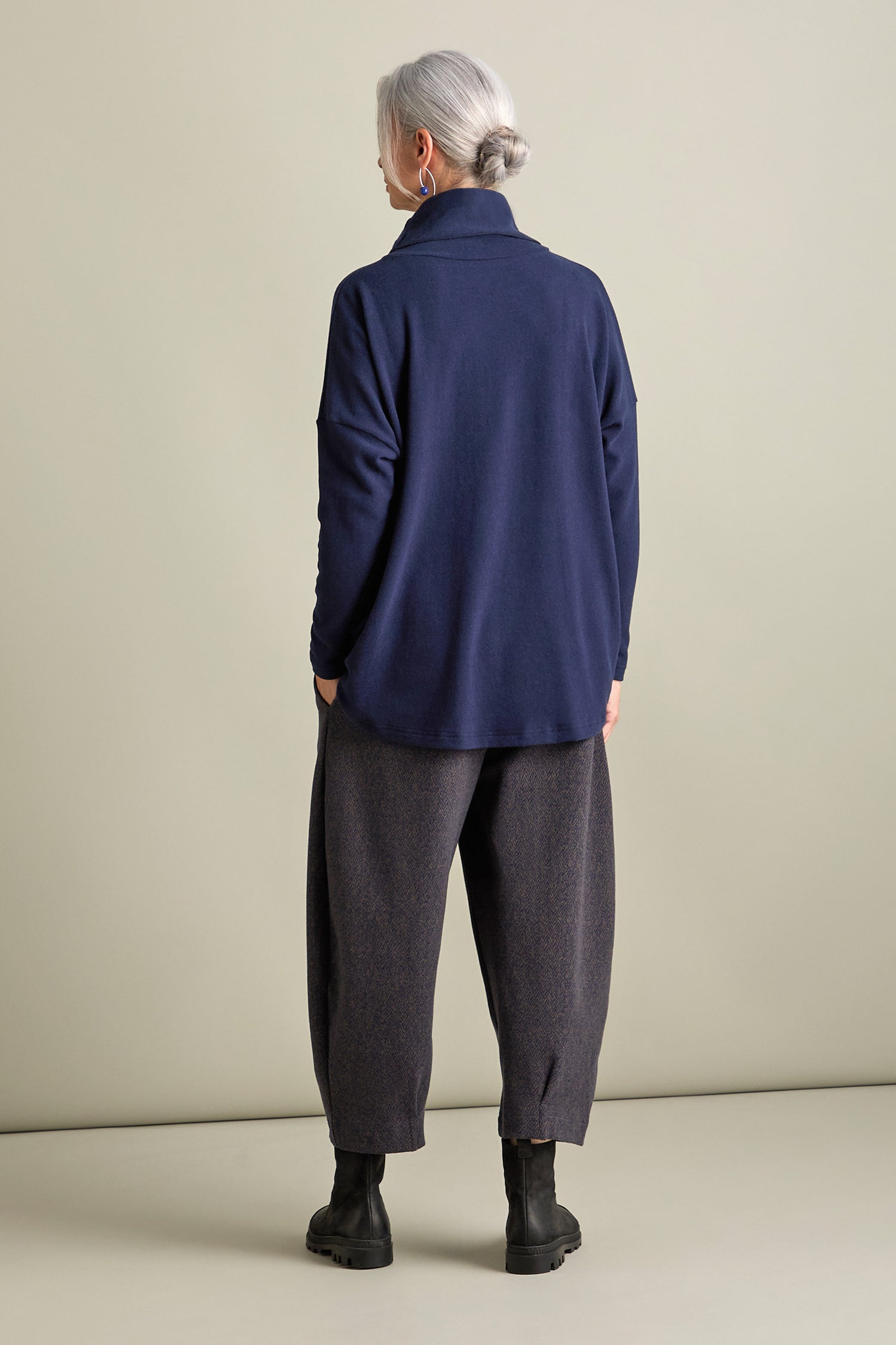 An individual with gray hair in a low bun is standing with their back to the camera, wearing the Bill Roll Neck Soft Jersey Top in navy blue, paired with gray loose-fitting pants and black boots, against a neutral background.