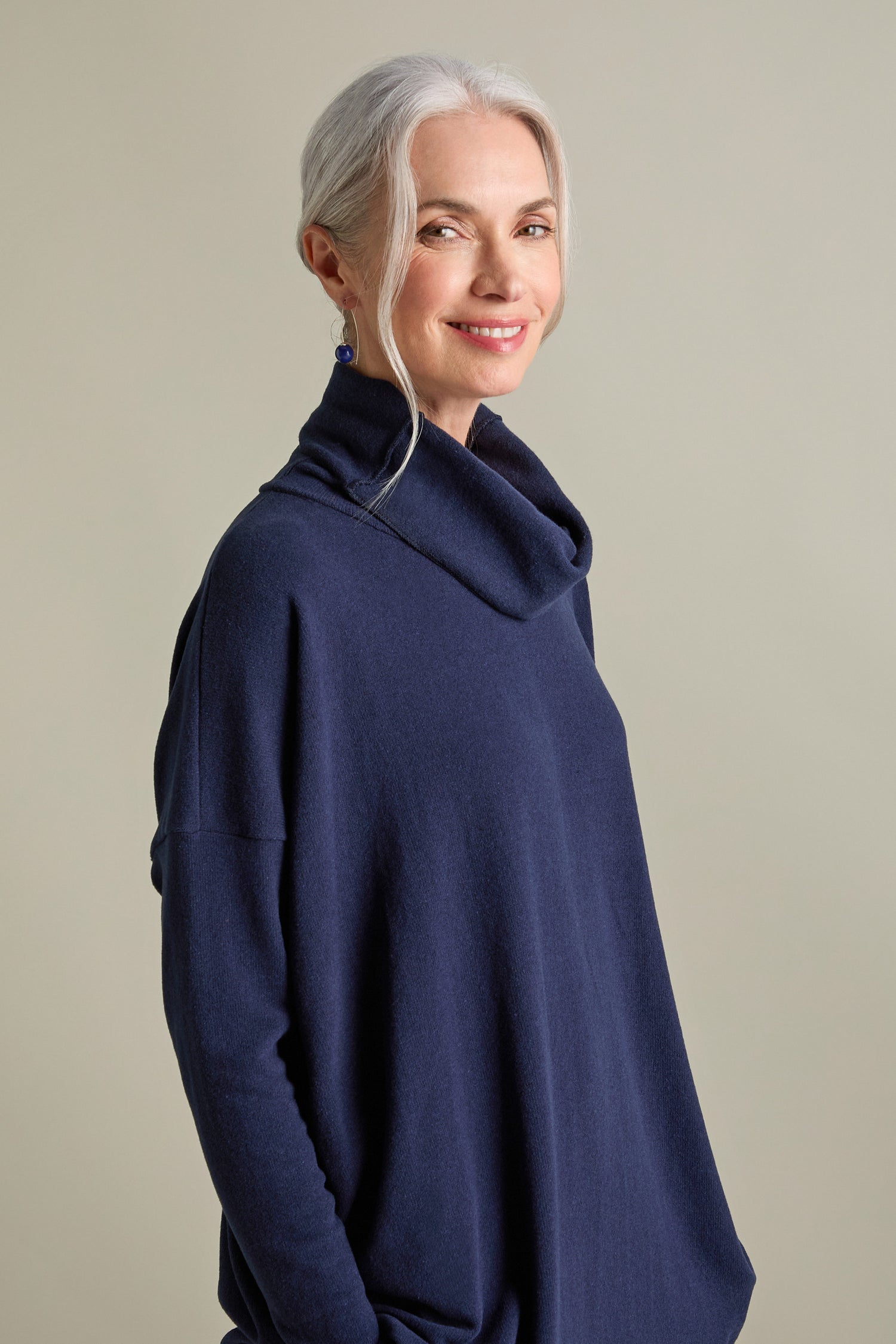 A woman with gray hair tied back is wearing a Bill Roll Neck Soft Jersey Top in dark blue and smiling slightly while looking at the camera. She has blue earrings and is standing against a plain background.