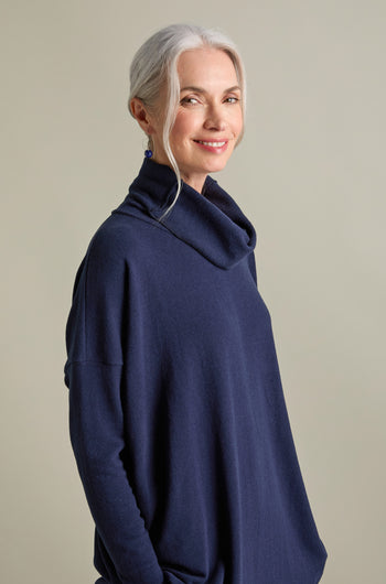 A woman with gray hair tied back is wearing a Bill Roll Neck Soft Jersey Top in dark blue and smiling slightly while looking at the camera. She has blue earrings and is standing against a plain background.