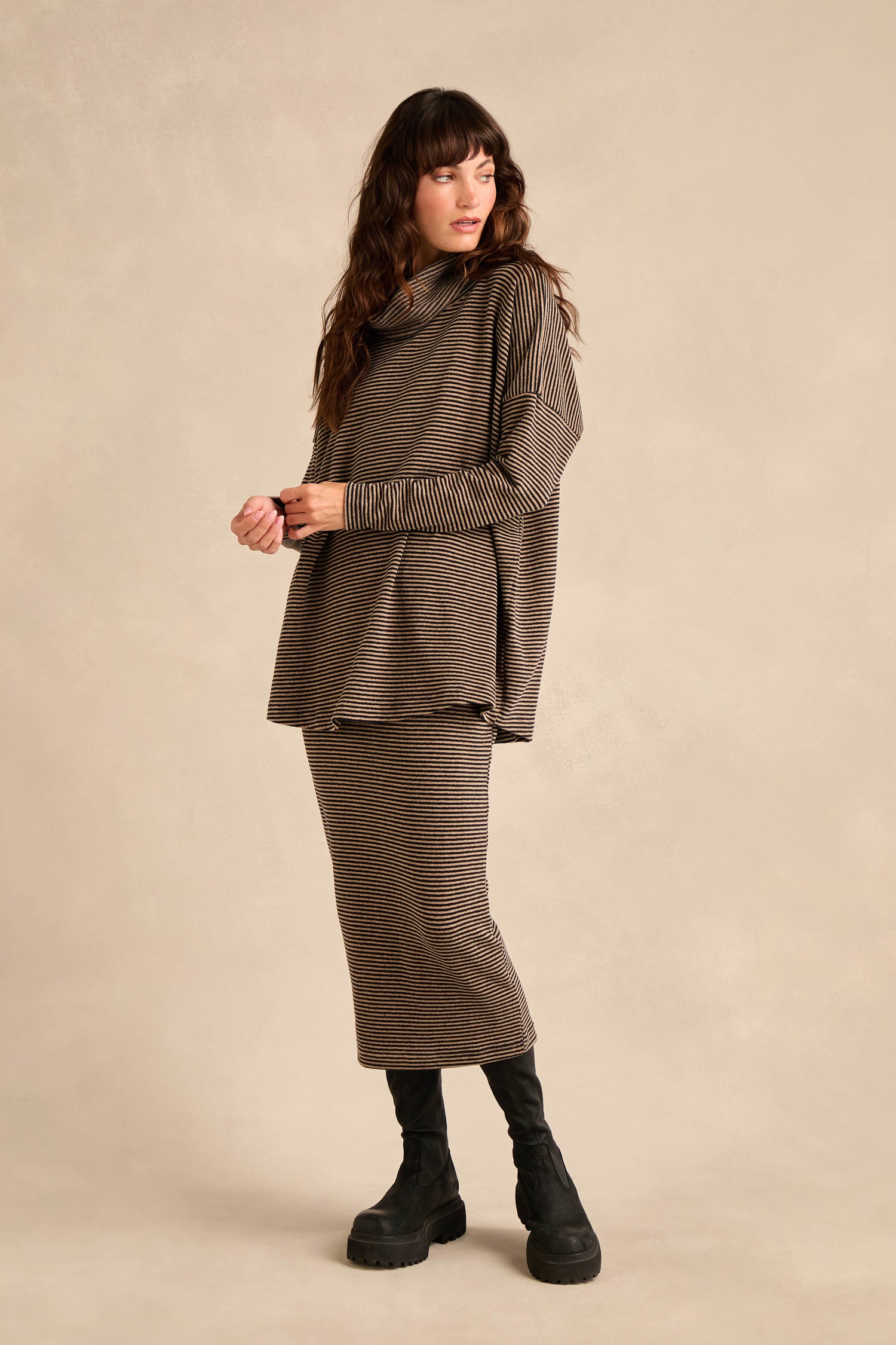 A person stands wearing the Roll Neck Narrow Stripe Fleece Tunic, a cosy long-sleeve piece featuring brown and black stripes, paired with a matching striped skirt and black boots against a neutral background.