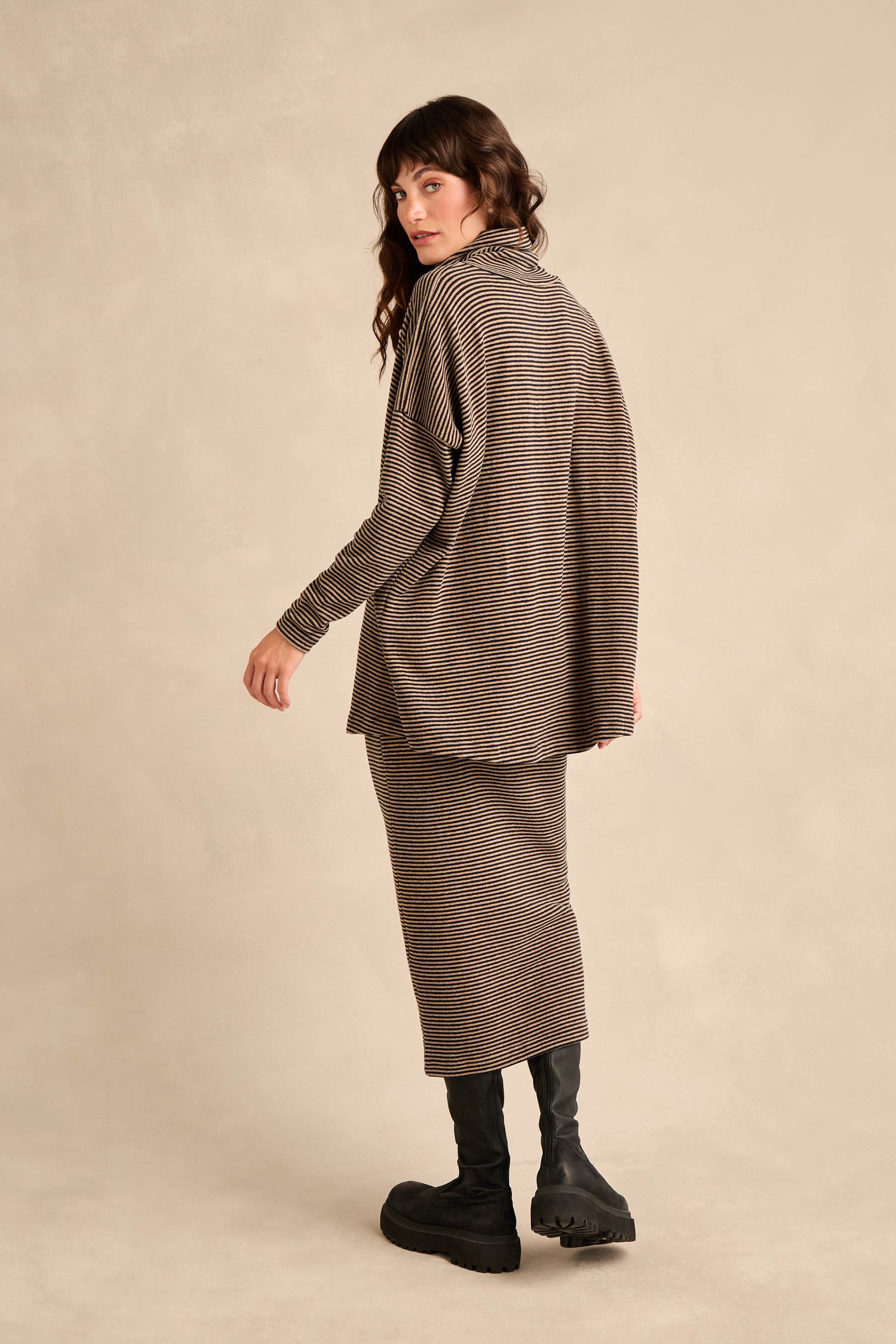A woman stands in profile against a neutral background, wearing the Roll Neck Narrow Stripe Fleece Tunic with brown stripes, a matching skirt, and black boots.
