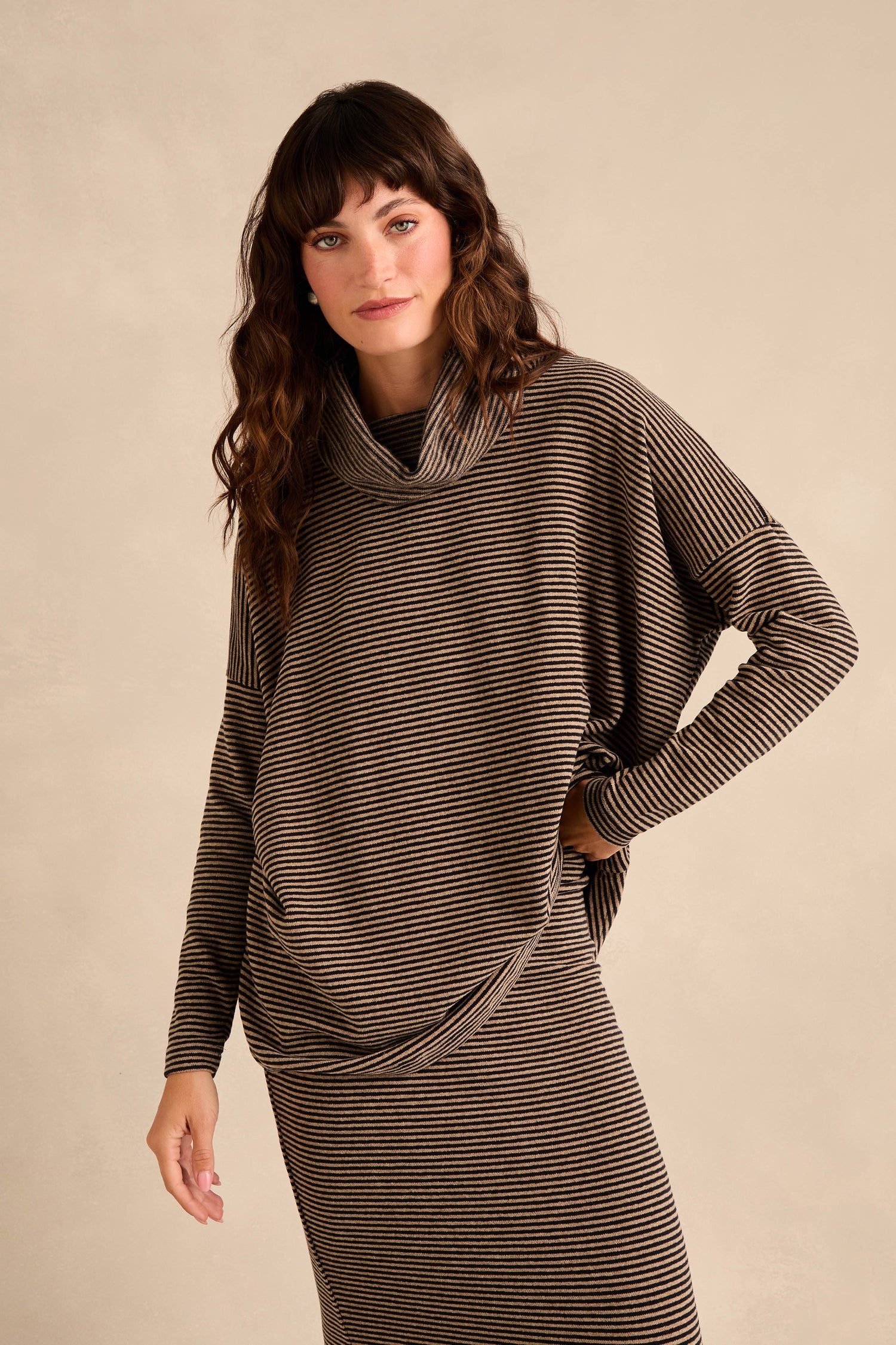 A person with wavy brown hair wears the Roll Neck Narrow Stripe Fleece Tunic in brown and black stripes, paired with a matching skirt, standing against a plain beige background.