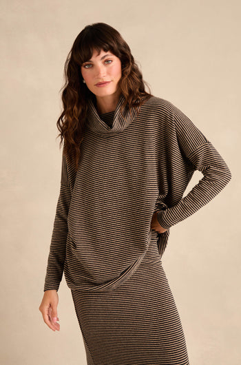 A person with wavy brown hair wears the Roll Neck Narrow Stripe Fleece Tunic in brown and black stripes, paired with a matching skirt, standing against a plain beige background.