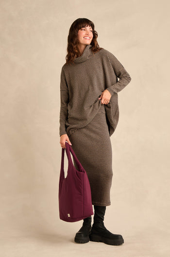 A woman wearing the Roll Neck Narrow Stripe Fleece Tunic paired with black boots and carrying a large purple tote bag stands against a beige background, smiling.