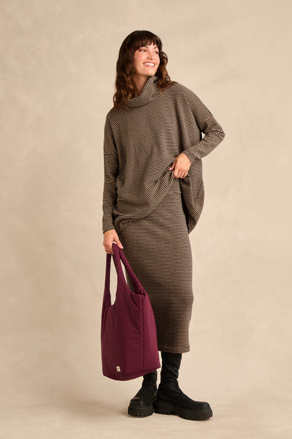 A woman wearing the Roll Neck Narrow Stripe Fleece Tunic paired with black boots and carrying a large purple tote bag stands against a beige background, smiling.