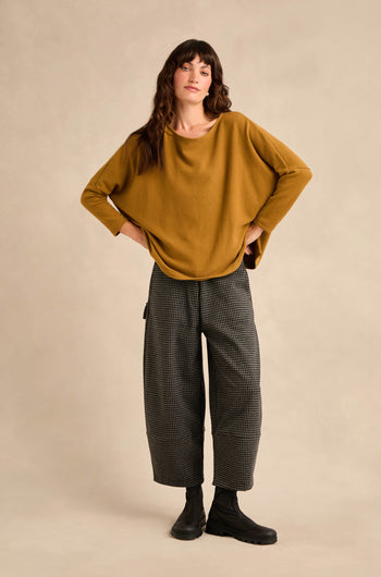 A person standing with hands on hips, wearing Mini Check Jersey Pocket Trousers featuring a cropped leg in soft cotton-blend jersey, paired with a mustard yellow sweater and black boots against a plain beige background.