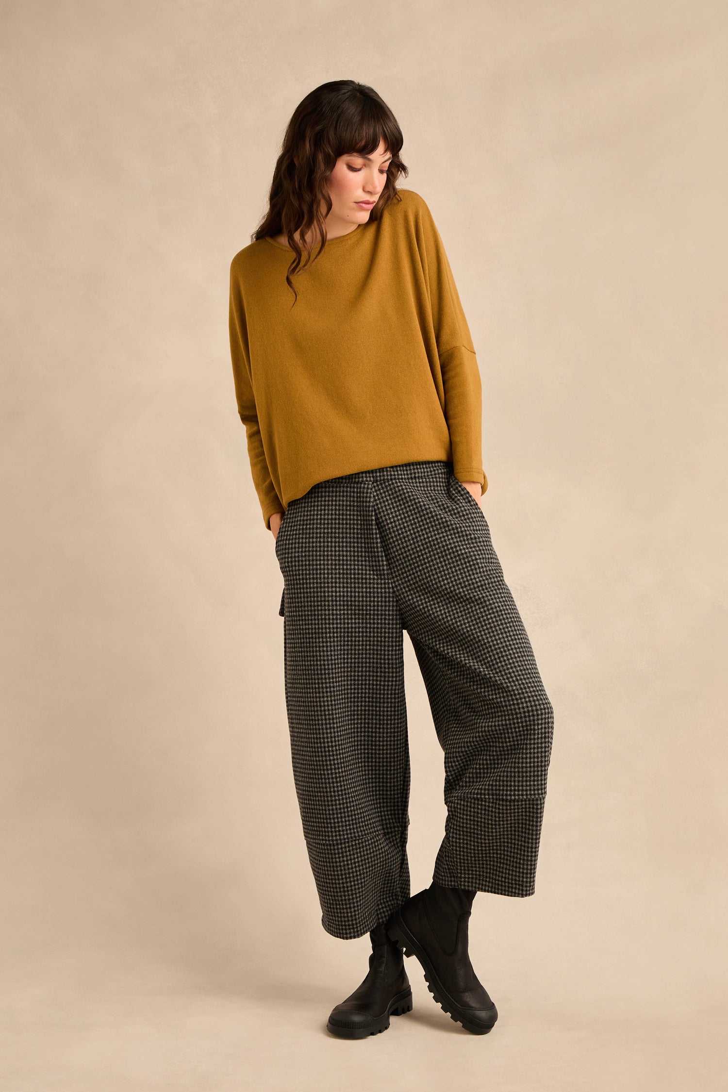 A person stands against a light background wearing a mustard yellow sweater, the Mini Check Jersey Pocket Trousers with a cropped leg, and black boots, looking down to the side with hands in pockets.
