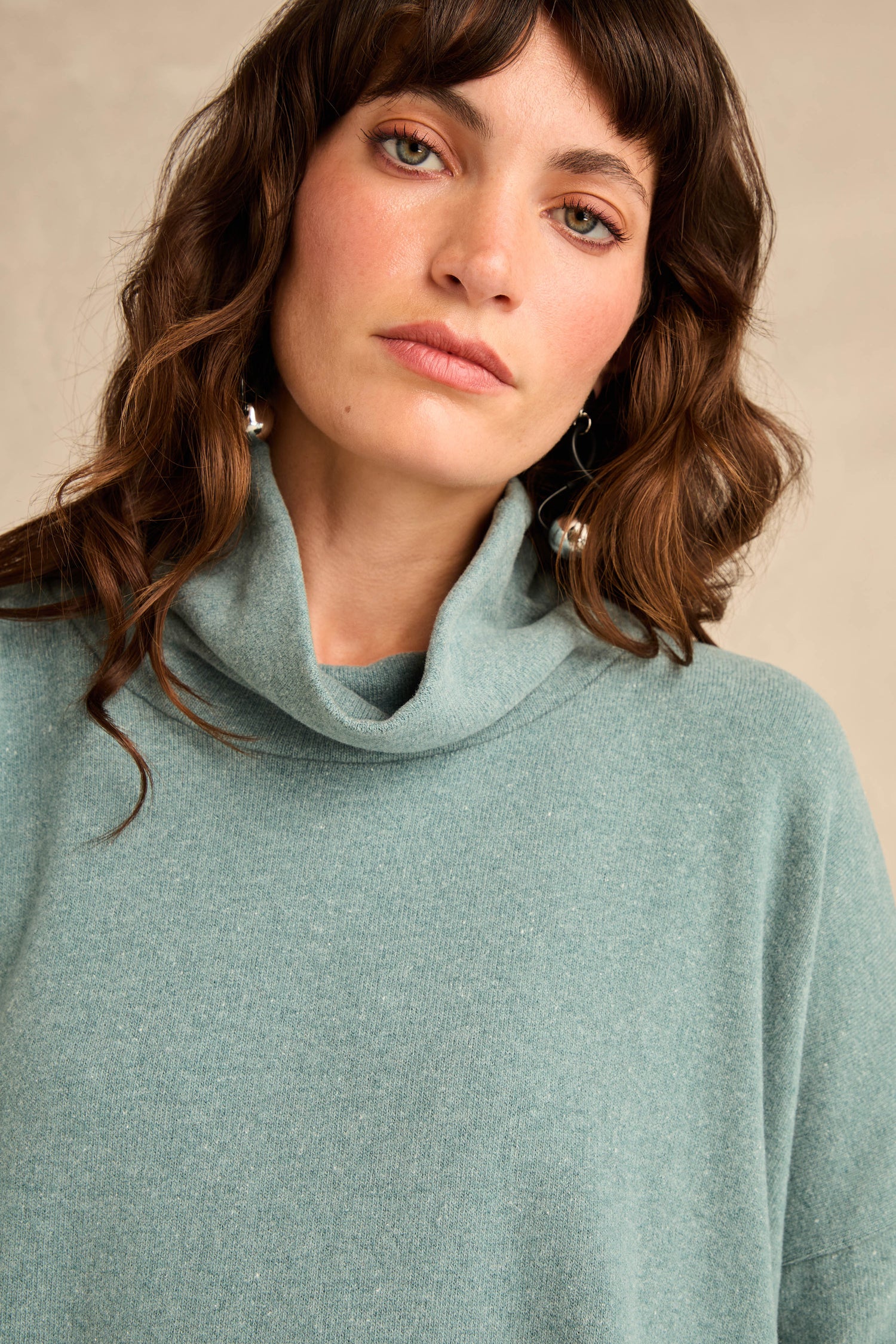 A woman with wavy brown hair is wearing a light teal Cowl Neck Soft Jersey Top and silver hoop earrings, looking at the camera with a neutral expression.