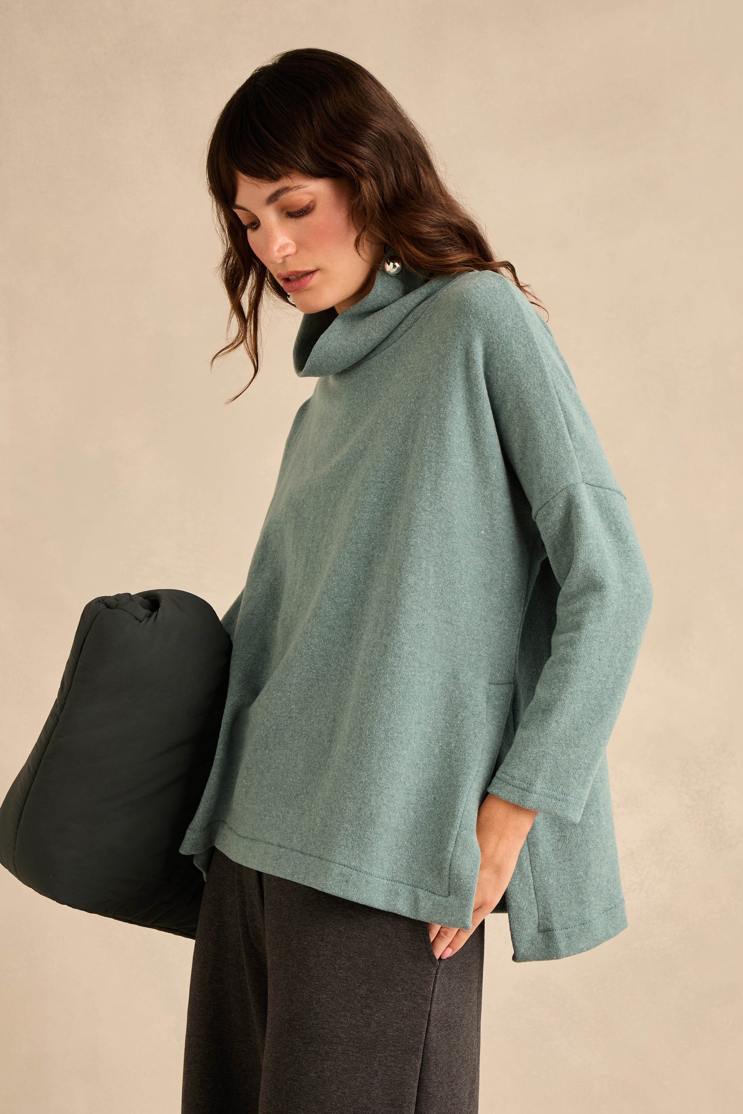 A woman wearing the Cowl Neck Soft Jersey Top in light teal paired with dark pants, holding a dark padded object against a beige background.