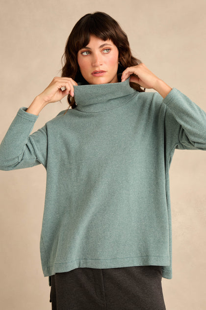 Wearing the Cowl Neck Soft Jersey Top in light teal, this person adjusts the collar of the loose-fitting, cotton-blend top while standing against a neutral background and pairing it with dark pants.