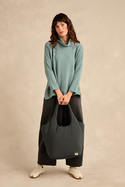 Woman standing against a beige background, wearing the Cowl Neck Soft Jersey Top in teal—a relaxed-fit piece crafted from cotton-blend jersey—paired with dark wide-leg pants and white sneakers, holding a large dark tote bag in front of her with both hands.