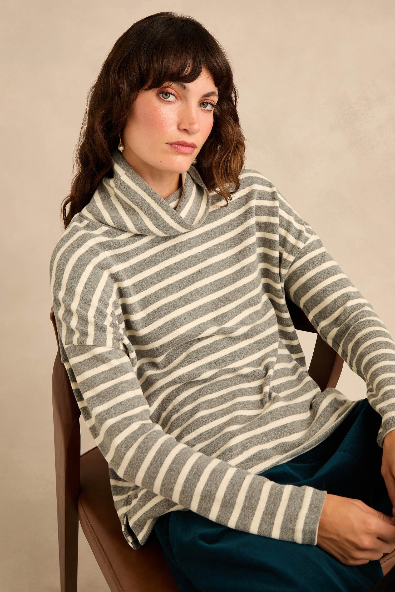 A woman with wavy brown hair sits on a chair against a neutral background, looking towards the camera with a relaxed expression. She is wearing the Roll Neck Silver Wide Stripe Jersey Top, featuring relaxed fit gray and white stripes in soft cotton-blend jersey.