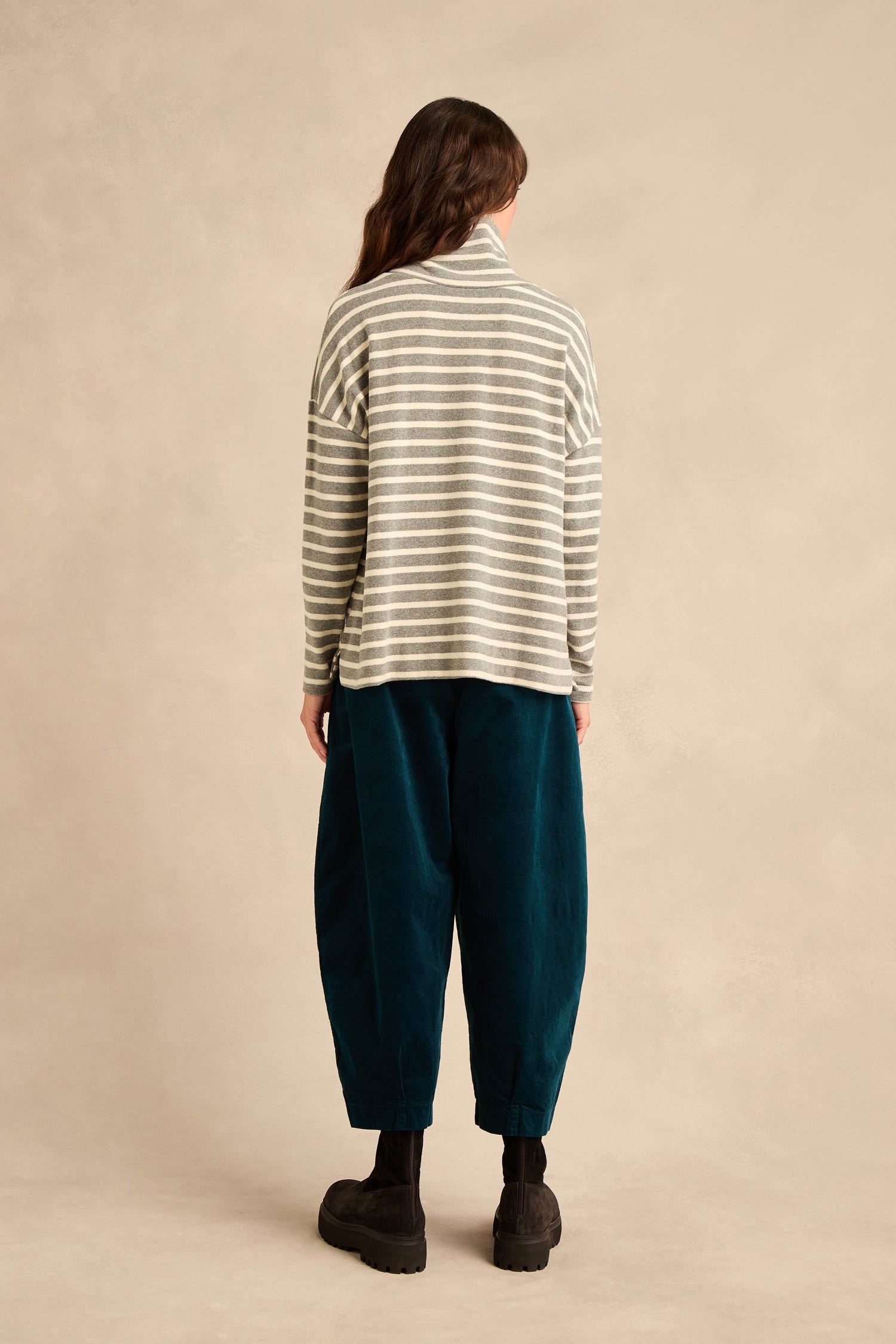 A person with long brown hair, seen from behind, wears the Roll Neck Silver Wide Stripe Jersey Top—crafted from cotton-blend jersey in a relaxed fit—paired with loose teal pants and black boots, standing against a beige background.