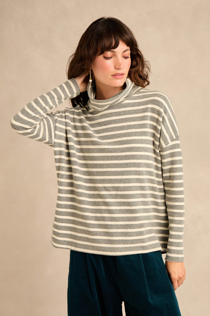 A woman wears the Roll Neck Silver Wide Stripe Jersey Top, featuring gray and white wide stripes in a relaxed fit crafted from soft cotton-blend jersey. She pairs it with teal pants and stands against a beige background, her hand gently touching her neck.