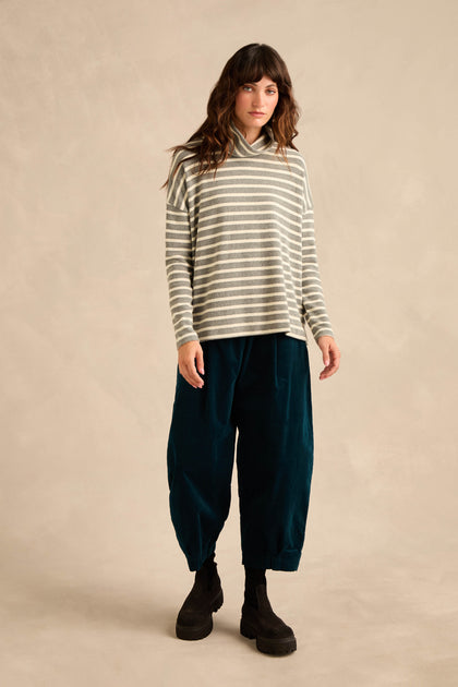 A woman stands against a plain beige background, wearing the Roll Neck Silver Wide Stripe Jersey Top—a relaxed fit made from soft cotton-blend jersey—paired with loose dark teal pants and black boots.