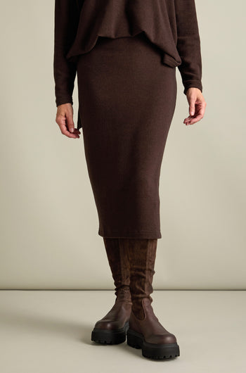 A person is standing, visible from shoulders down, wearing a brown sweater, a Lilla Soft Jersey Tube Skirt from the Autumn/Winter '24 collection, and dark brown knee-high boots with platform soles. The background is plain and neutral.