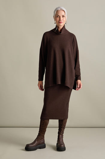 A woman with gray hair stands against a plain background wearing a long brown sweater, a matching Lilla Soft Jersey Tube Skirt from Mama b, and brown knee-high boots, embodying the cozy style of Autumn/Winter '24.