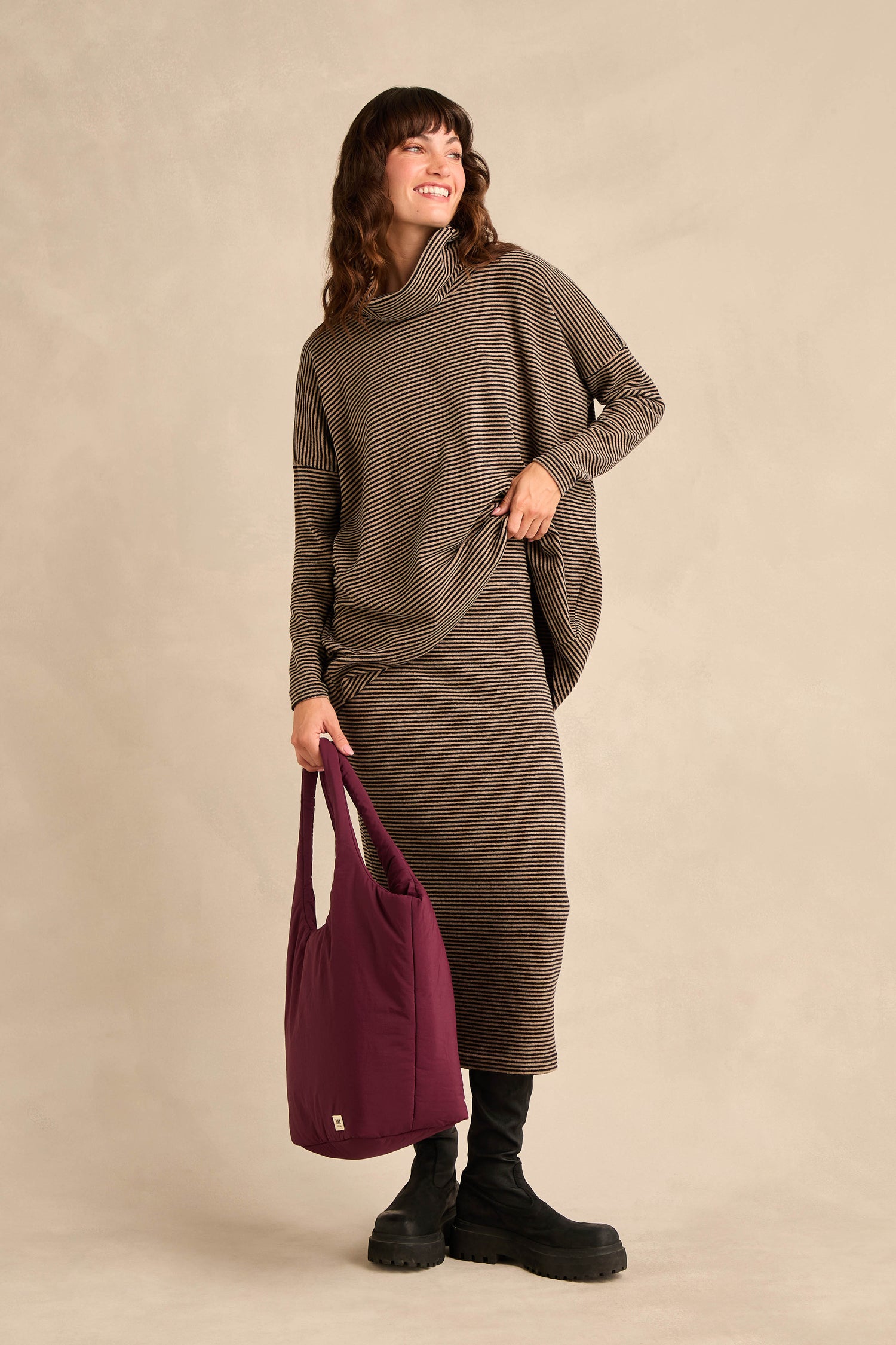 A woman stands smiling against a neutral background, wearing a Narrow Stripe Jersey Tube Skirt with a matching long-sleeve top, black boots, and holding a purple tote bag.
