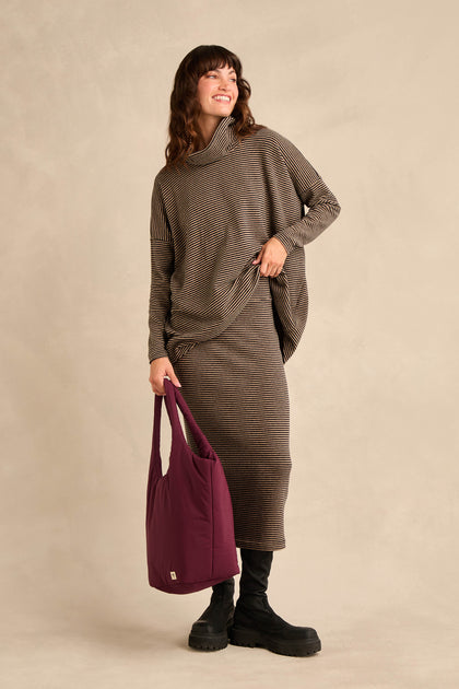 A woman stands smiling against a neutral background, wearing a Narrow Stripe Jersey Tube Skirt with a matching long-sleeve top, black boots, and holding a purple tote bag.