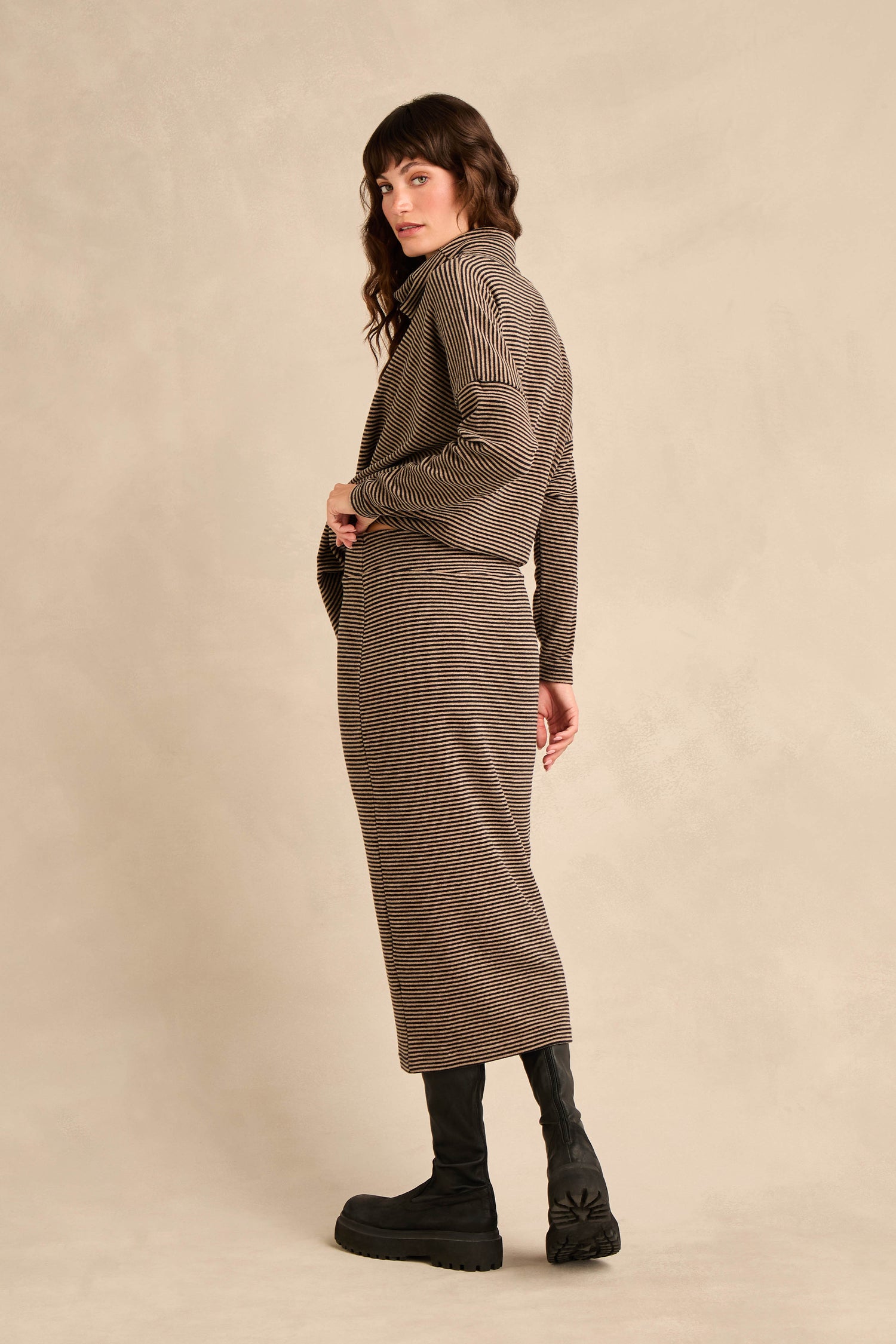 A woman poses against a beige background, looking over her shoulder in the Narrow Stripe Jersey Tube Skirt and matching long-sleeve top, styled with black boots.