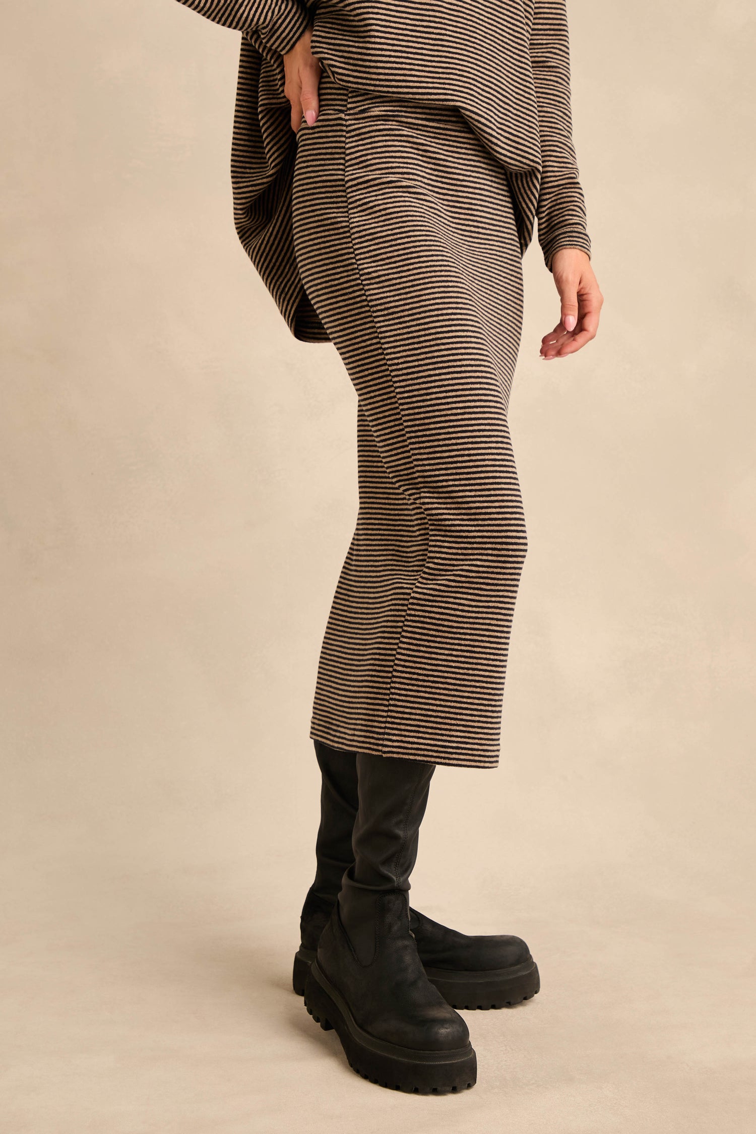 A person wearing the Narrow Stripe Jersey Tube Skirt in brown and black narrow stripes, paired with a long matching top and black boots, stands against a plain beige background.