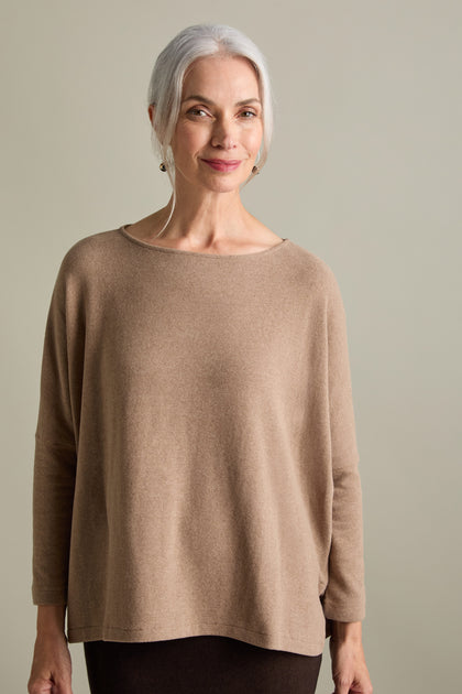 A woman with gray hair stands against a plain background, smiling gently while wearing a long-sleeved, beige Martis Scoop Neck Soft Jersey Top made in Italy.