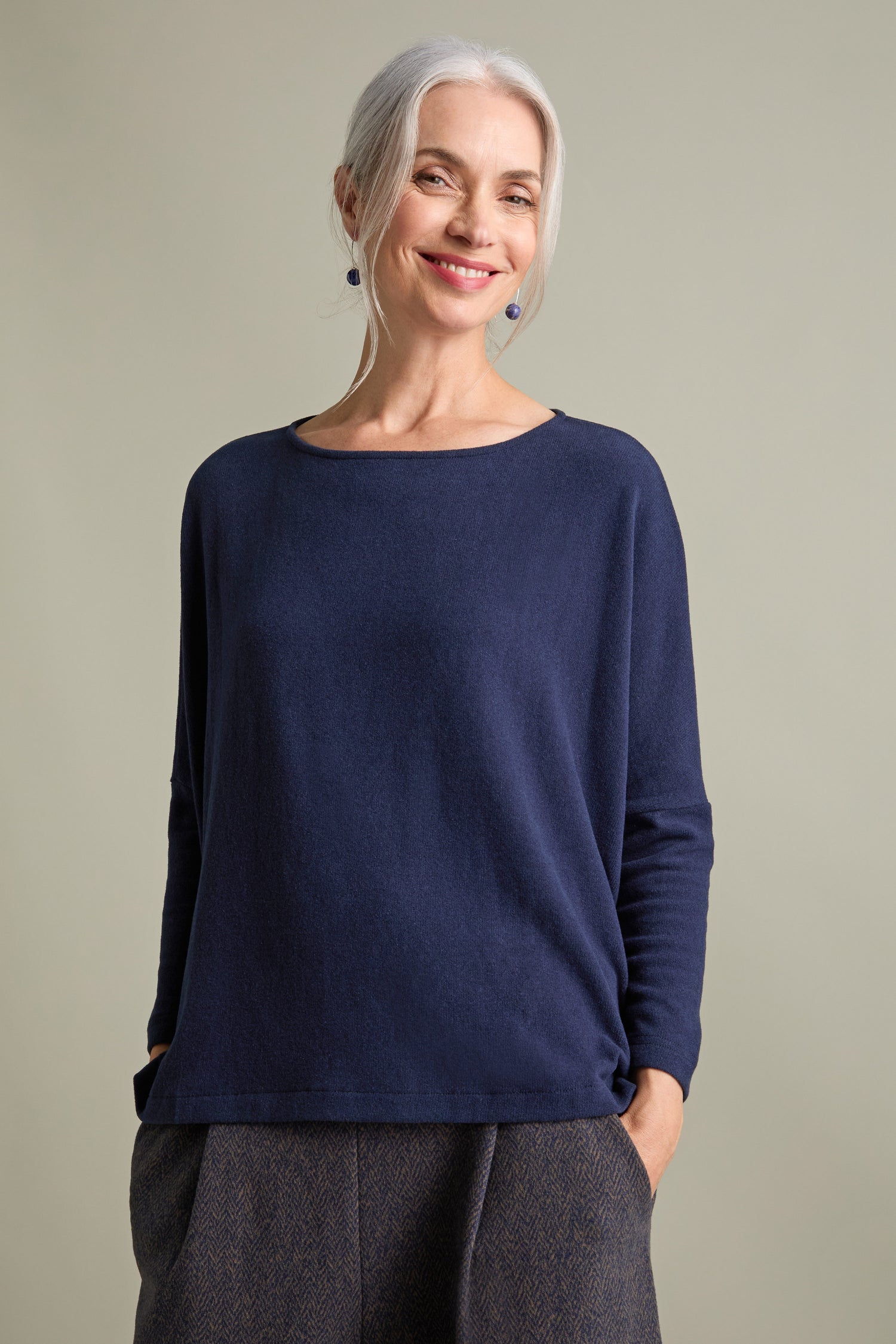 A woman with grey hair smiles while wearing a Martis Scoop Neck Soft Jersey Top made in Italy, paired with patterned trousers. She has her hands in her pockets.