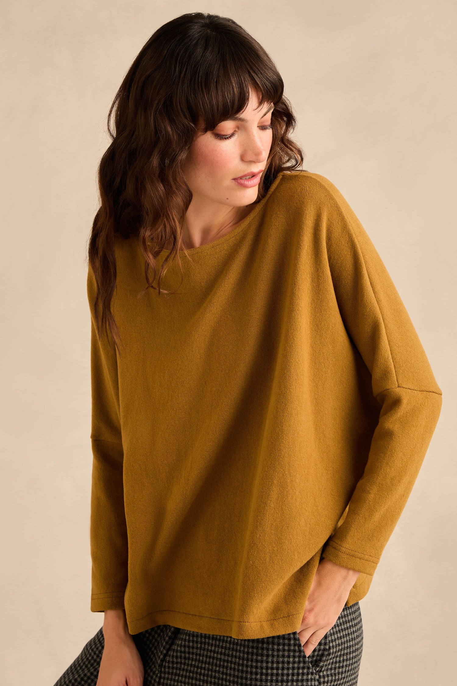 A woman wears the Round Neck Soft Jersey Top in a loose-fitting mustard yellow style paired with plaid pants. Standing against a neutral background and looking down to the side, she highlights the relaxed, everyday shape of this cotton-blend jersey piece for effortless comfort.