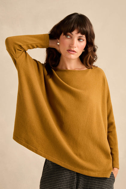 A woman wearing a loose-fitting, long-sleeve Round Neck Soft Jersey Top in mustard and checked pants stands with one hand touching her ear against a neutral background.
