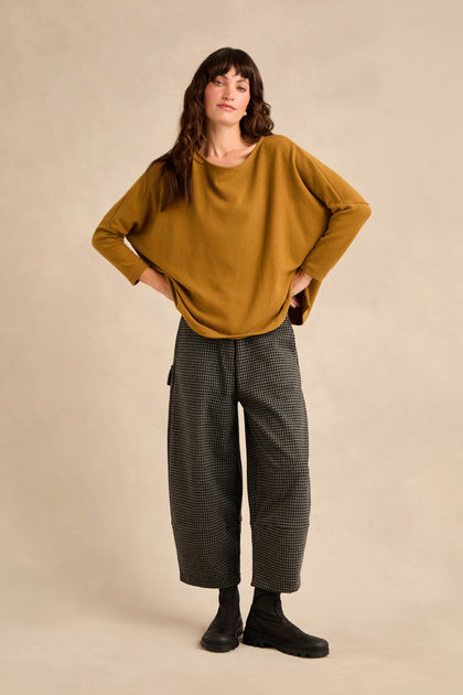 A woman stands with her hands on her hips, wearing the Round Neck Soft Jersey Top in a loose mustard yellow style, paired with wide gray checkered pants and black ankle boots, set against a plain beige background.