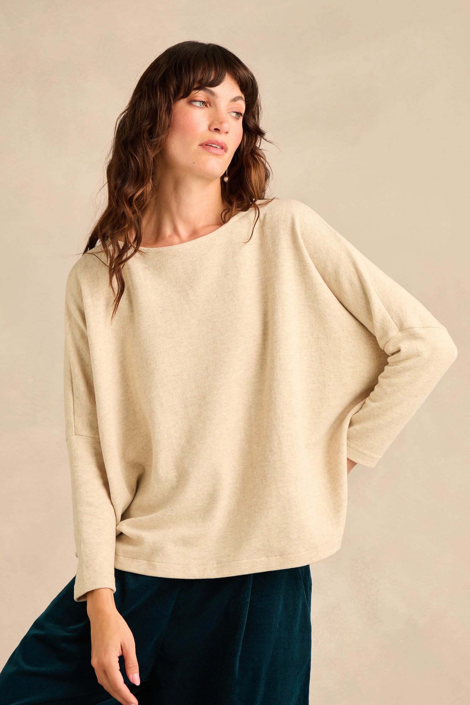 Person wearing the Round Neck Soft Jersey Top in beige—a loose-fitting cotton-blend jersey with long sleeves—paired with dark pants, posing with one hand on their hip against a plain background. The soft top features a relaxed, everyday silhouette for effortless style.
