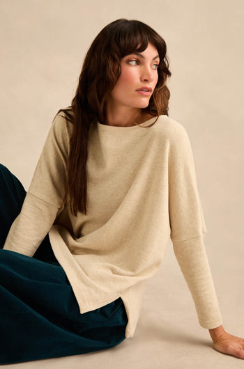 A woman with long brown hair wears the Round Neck Soft Jersey Top—a loose, relaxed everyday cotton-blend jersey sweater—paired with dark pants while sitting on the floor and looking to her left against a neutral background.
