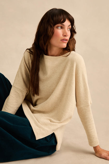 A woman with long brown hair wears the Round Neck Soft Jersey Top—a loose, relaxed everyday cotton-blend jersey sweater—paired with dark pants while sitting on the floor and looking to her left against a neutral background.