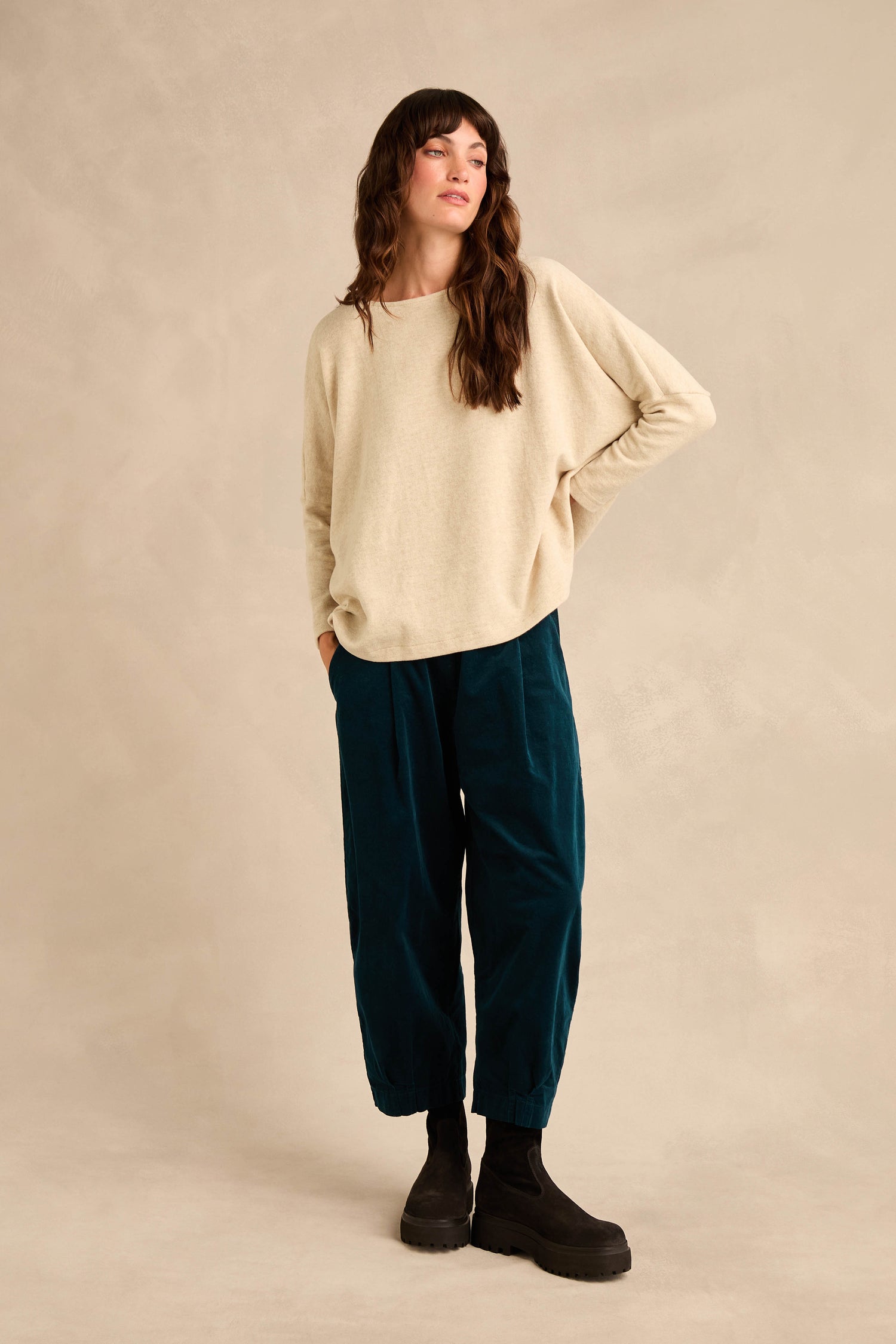 A woman stands against a neutral background wearing the Round Neck Soft Jersey Top—a loose beige cotton-blend jersey sweater—paired with teal pants and black boots. With one hand in her pocket and her head turned slightly to the side, she showcases the top’s relaxed everyday shape that offers effortless style.