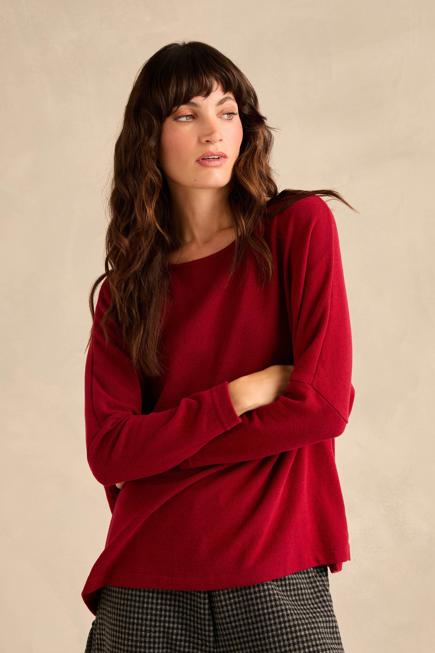A woman with long brown hair stands against a neutral background, arms crossed, wearing the Round Neck Soft Jersey Top in red and patterned pants.