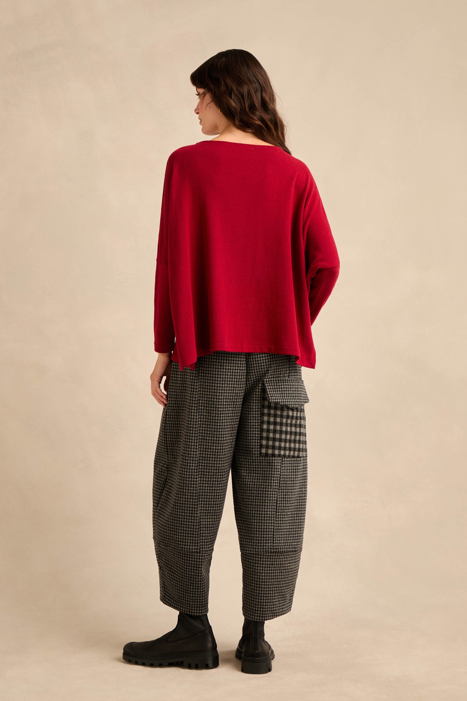 A person stands on a light background, facing away. They are wearing the Round Neck Soft Jersey Top in red, which has a relaxed, loose fit, paired with gray checkered pants with patch pockets and black boots.