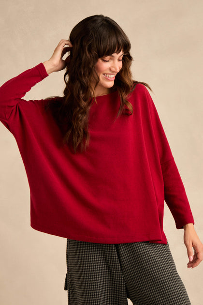 A woman wearing the Round Neck Soft Jersey Top in red—a loose-fitting, long-sleeve cotton-blend jersey with a relaxed everyday silhouette—paired with checked wide-leg pants, stands against a plain beige background, looking down and smiling.
